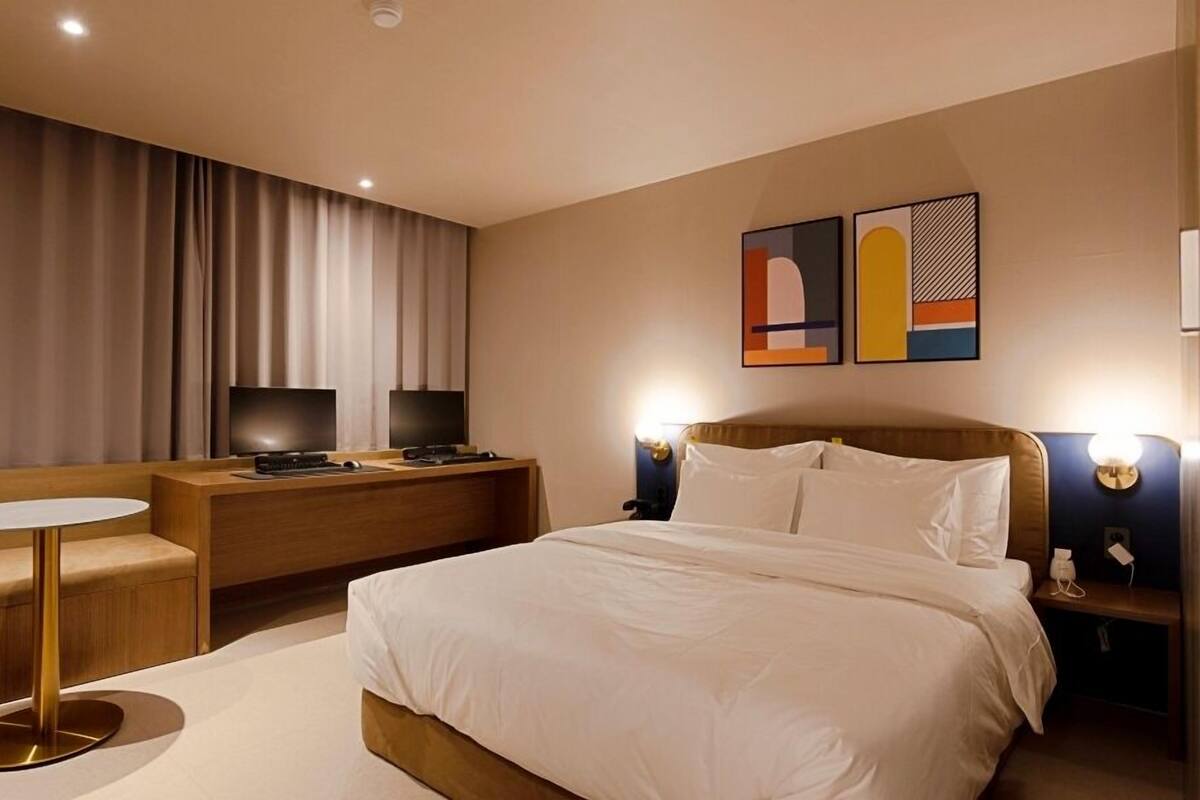 A hotel room with a bed, a desk, a chair, and two wall-mounted televisions.