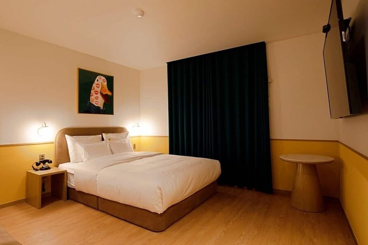 A hotel room with a bed, bedside lamps, a small table, a painting on the wall, and a TV mounted on the wall.