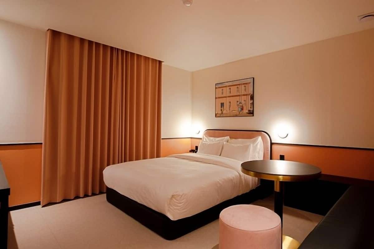 A hotel room with a bed, a round side table, a small ottoman, a painting on the wall, and a window with curtains.