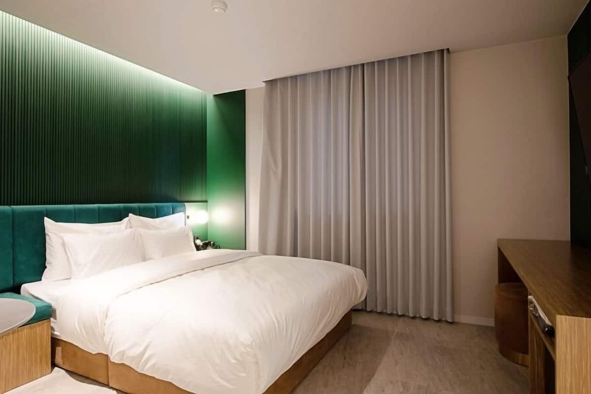 A modern hotel room with a large bed, a green accent wall, a wooden desk, and a large window with curtains.