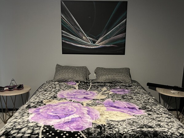 Een slaapkamer met een bed, twee kussens, een nachtkastje en een kunstwerk aan de muur.