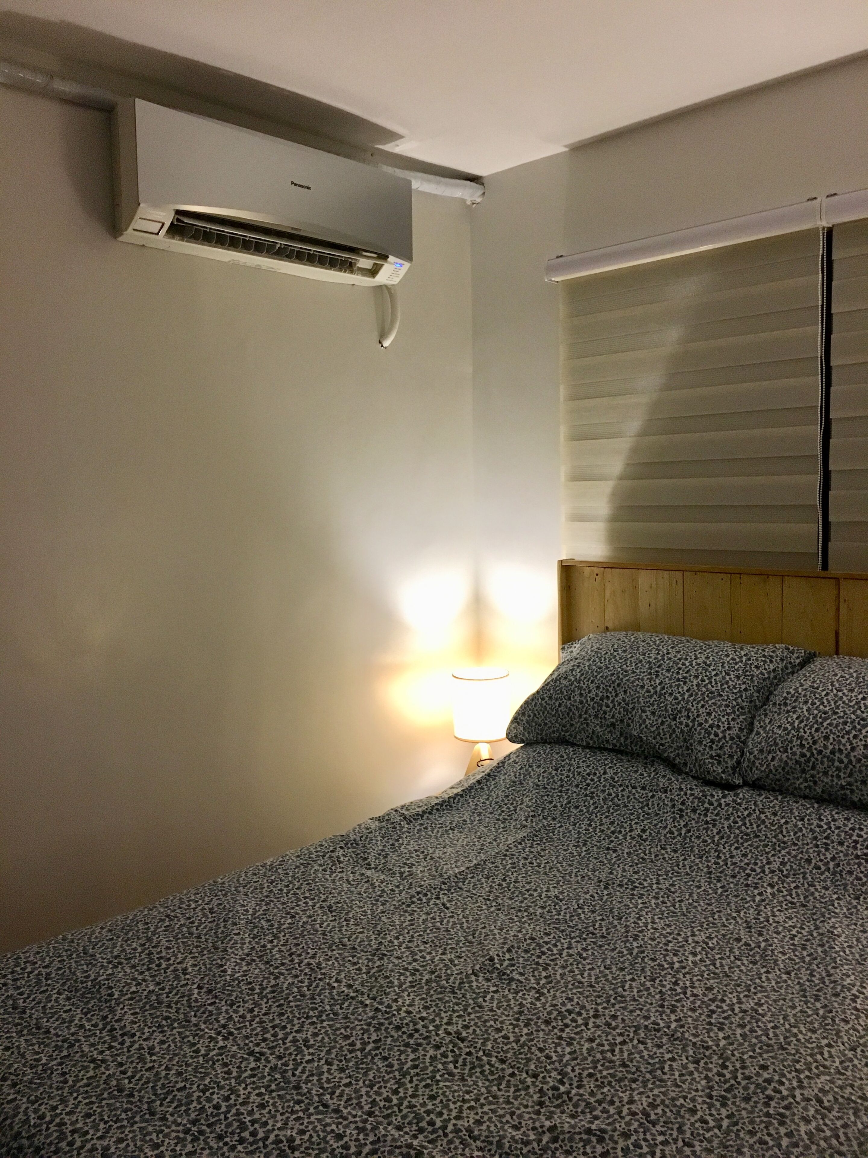 Une chambre d’hôtel équipée d’un lit, d’un appareil de climatisation, d’une fenêtre avec des stores et d’une lampe de chevet.