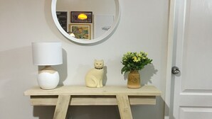 Una habitación con una puerta blanca, un espejo redondo, una pequeña mesa blanca con una lámpara, una figurita de gato, un florero con flores y cestas tejidas en estantes.