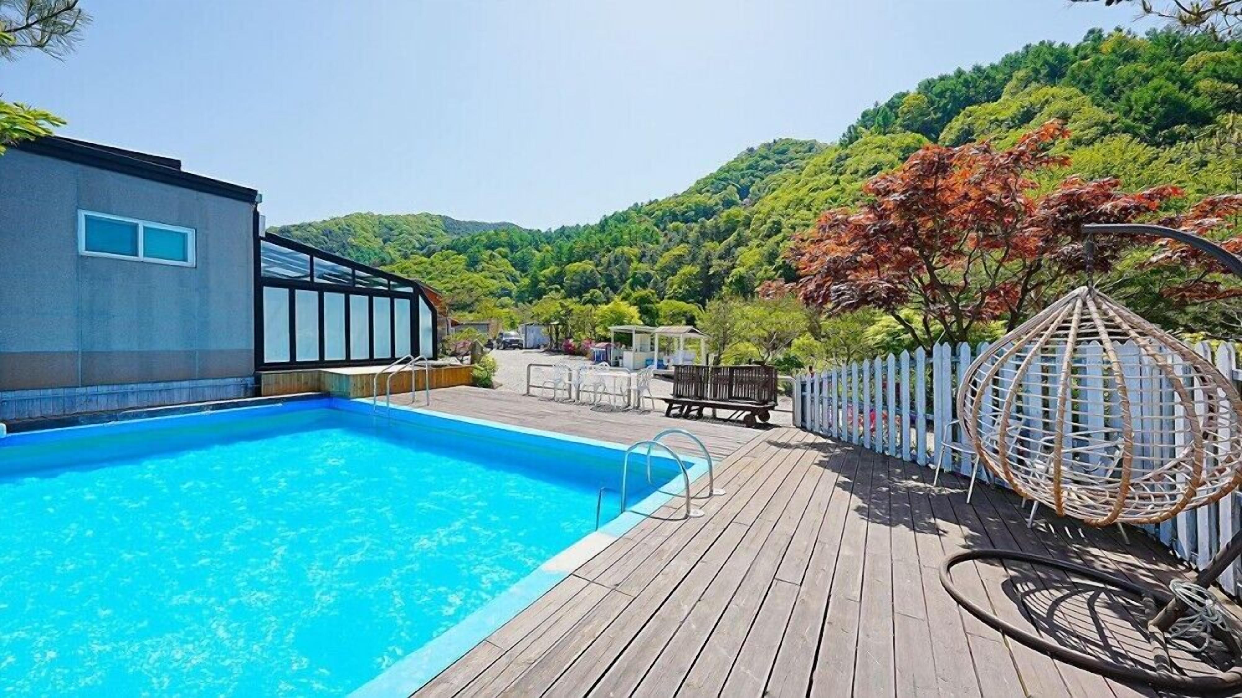 Una piscina con muelle de madera, zona de estar cubierta y vistas a un paisaje boscoso.