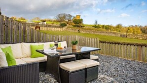 An outdoor seating area with wicker furniture and a table, overlooking a stone wall and a grassy field with a castle-like structure in the distance. - Yew Tree Cottage | Farm Stay | Sleeps 4 (West Ayton)