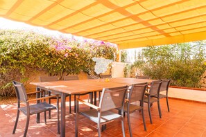 An outdoor patio with a wooden table and chairs, surrounded by lush greenery and flowering plants.