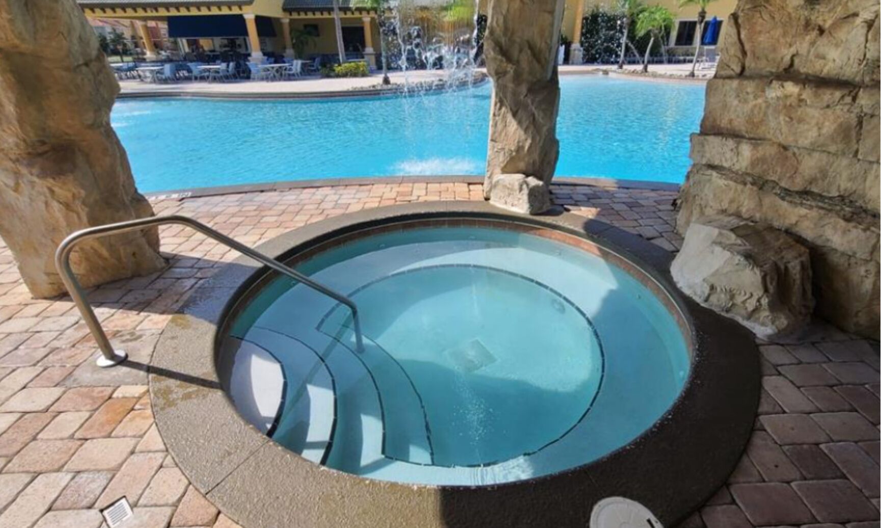 A hot tub with a metal railing, surrounded by stone pillars, with a pool and lounge area in the background.