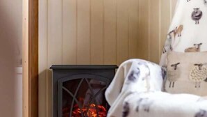 A cozy room with a wood-burning stove, a chair with a blanket, and a wooden wall. - Shepherds Hut | Cosy Farm Stay | Sleeps 2 (West Ayton)