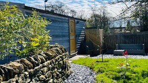 A rustic cabin with a stone wall, a wooden door, and a small garden area with gravel and grass. - Shepherds Hut | Cosy Farm Stay | Sleeps 2 (West Ayton)