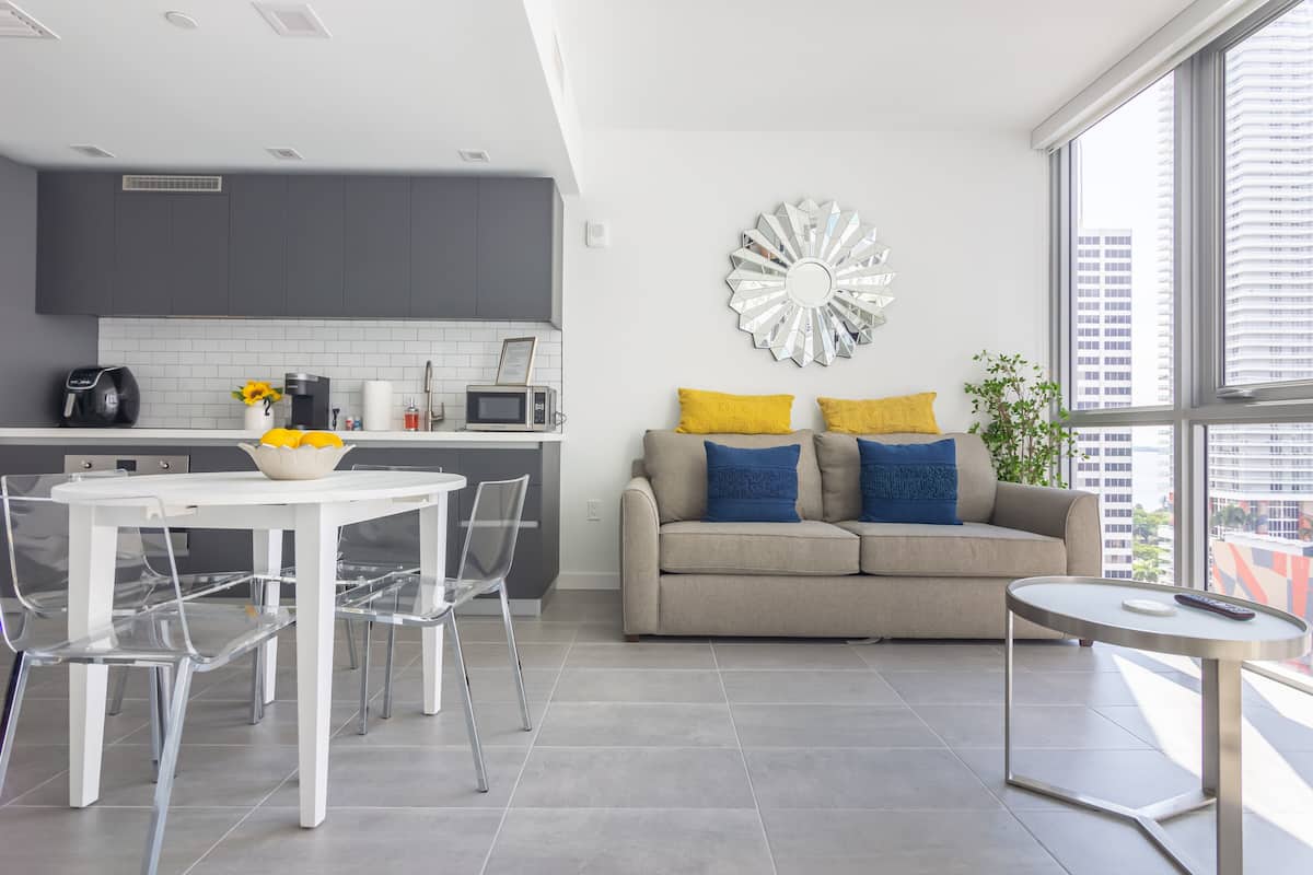 A modern kitchen with grey cabinets, a white dining table with clear chairs, and a grey tiled floor.