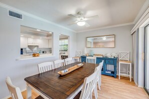A dining area with a wooden table, white chairs, a ceiling fan, and a kitchen in the background.
