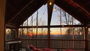Uma área de estar aconchegante ao ar livre, com móveis de vime e almofadas vermelhas, oferece vista panorâmica para um pôr do sol através de grandes janelas.