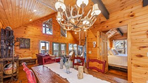 Interior de cabana de toras com uma mesa de jantar, cadeiras vermelhas, um candelabro e uma janela grande.