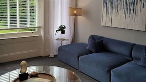 A modern living room with a blue sofa, a round glass coffee table, a floor lamp, and a large abstract painting on the wall. - Crescent View | Central & Quiet | Sleeps 4 (North Yorkshire)