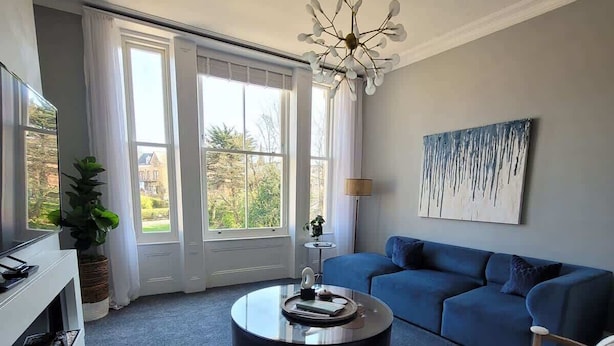 A modern living room with a blue sofa, a round coffee table, a white armchair, and a unique ceiling light fixture. - Crescent View | Central & Quiet | Sleeps 4 (North Yorkshire)