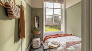 A bedroom with a bed, a bedside table with books and a lamp, a window with a view of a garden, and a hanging basket on the wall. - Crescent View | Central & Quiet | Sleeps 4 (North Yorkshire)