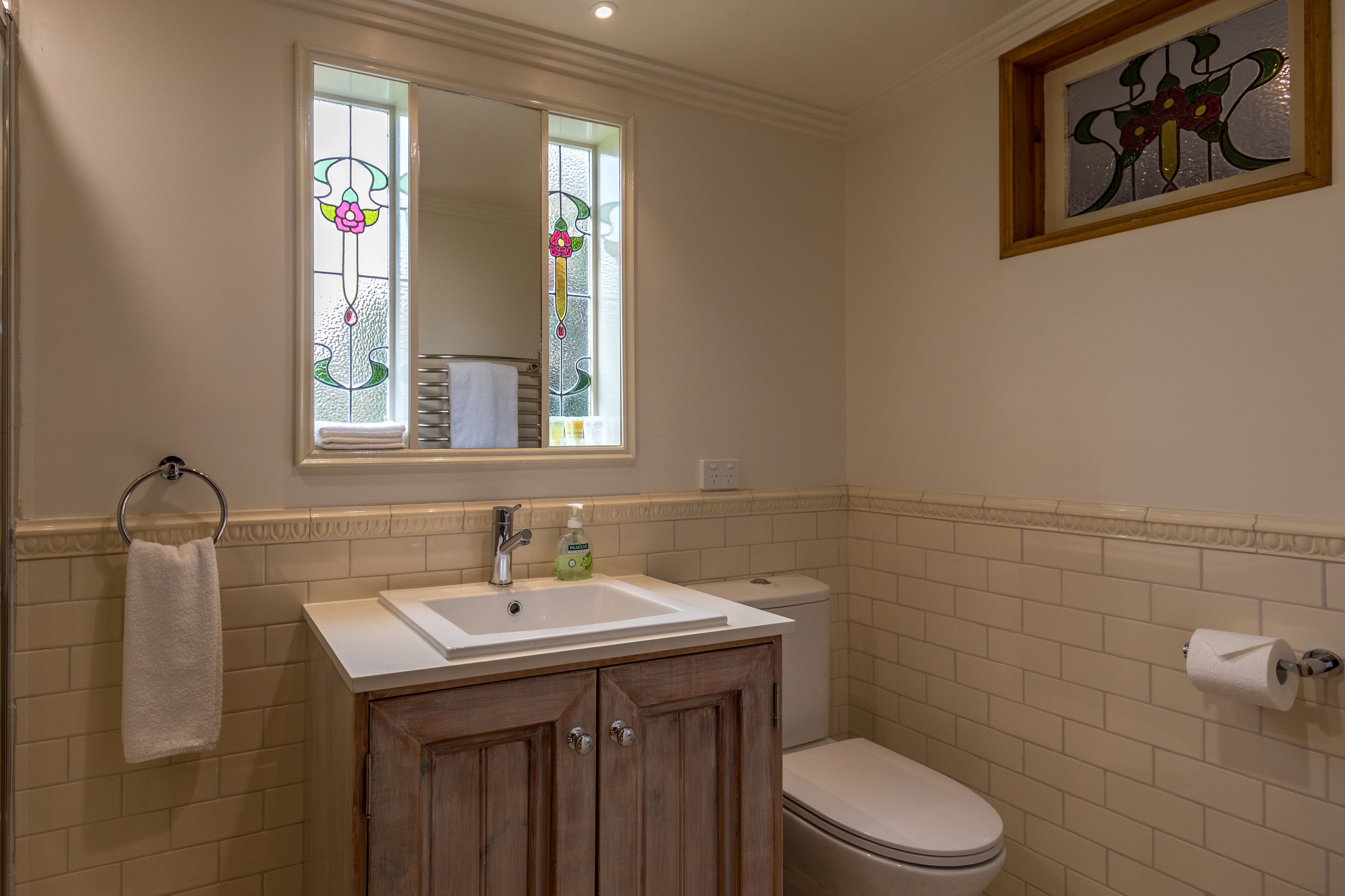A bathroom with a white sink, wooden cabinets, a toilet, and a stained glass window above the mirror.