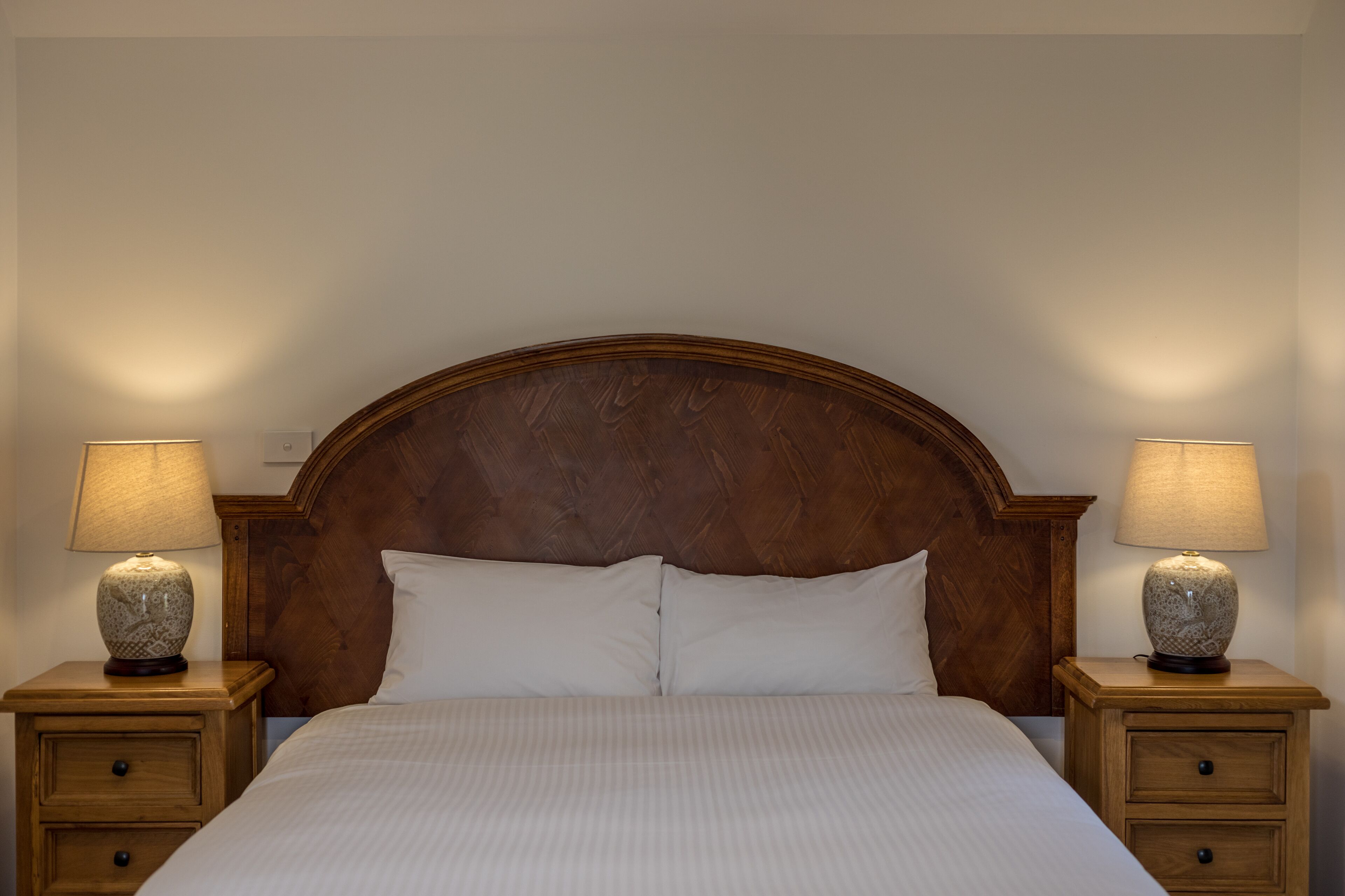Un lit bien fait, avec une tête de lit en bois, deux tables de chevet avec des lampes et un mur sobre.