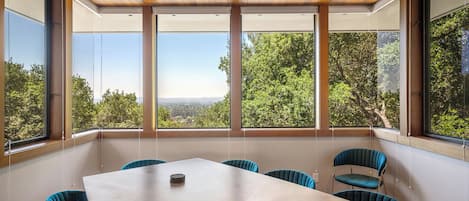 A conference room with a large table and six chairs, large windows with a view of trees and hills, wooden ceiling with recessed lighting, and a white wall.