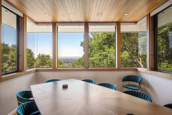 A conference room with a large table and six chairs, large windows with a view of trees and hills, wooden ceiling with recessed lighting, and a white wall.