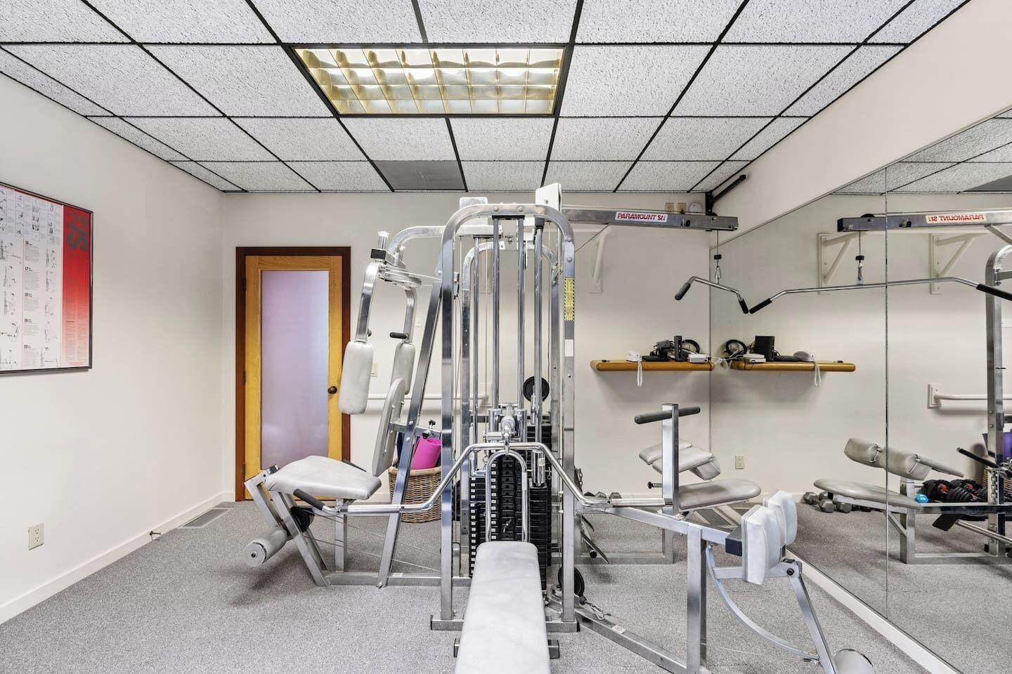 Un gimnasio con diversas máquinas de ejercicio, una puerta y un estante con objetos en la pared.