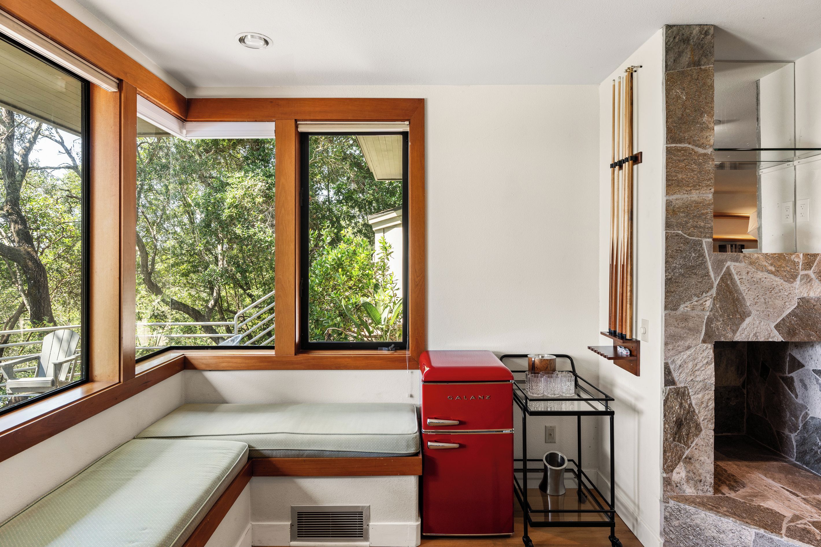 A compact kitchen with a built-in stove, a red refrigerator, and a window seat with a view of trees.