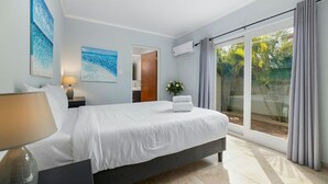 A modern hotel room with a large bed, a bedside table with a lamp, a window with curtains, and a view of a garden. - Great Cozy, Comfortable & Central 2BR 2BA w/ Pool (Noord)
