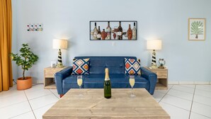 A living room with a blue sofa, two side tables with lamps, a wooden coffee table with a champagne bottle and glasses, a wall-mounted art piece of wine bottles, and a potted plant. - Spacious 1-bed w/ courtyard near Palm Beach (Palm Beach)