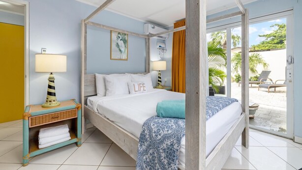 A four-poster bed with a white and blue patterned bedspread, two white pillows, and a single teal pillow. There is a wicker side table with a lamp and a small shelf with towels. A framed picture hangs on the wall above the bed. - Spacious 1-bed w/ courtyard near Palm Beach (Palm Beach)