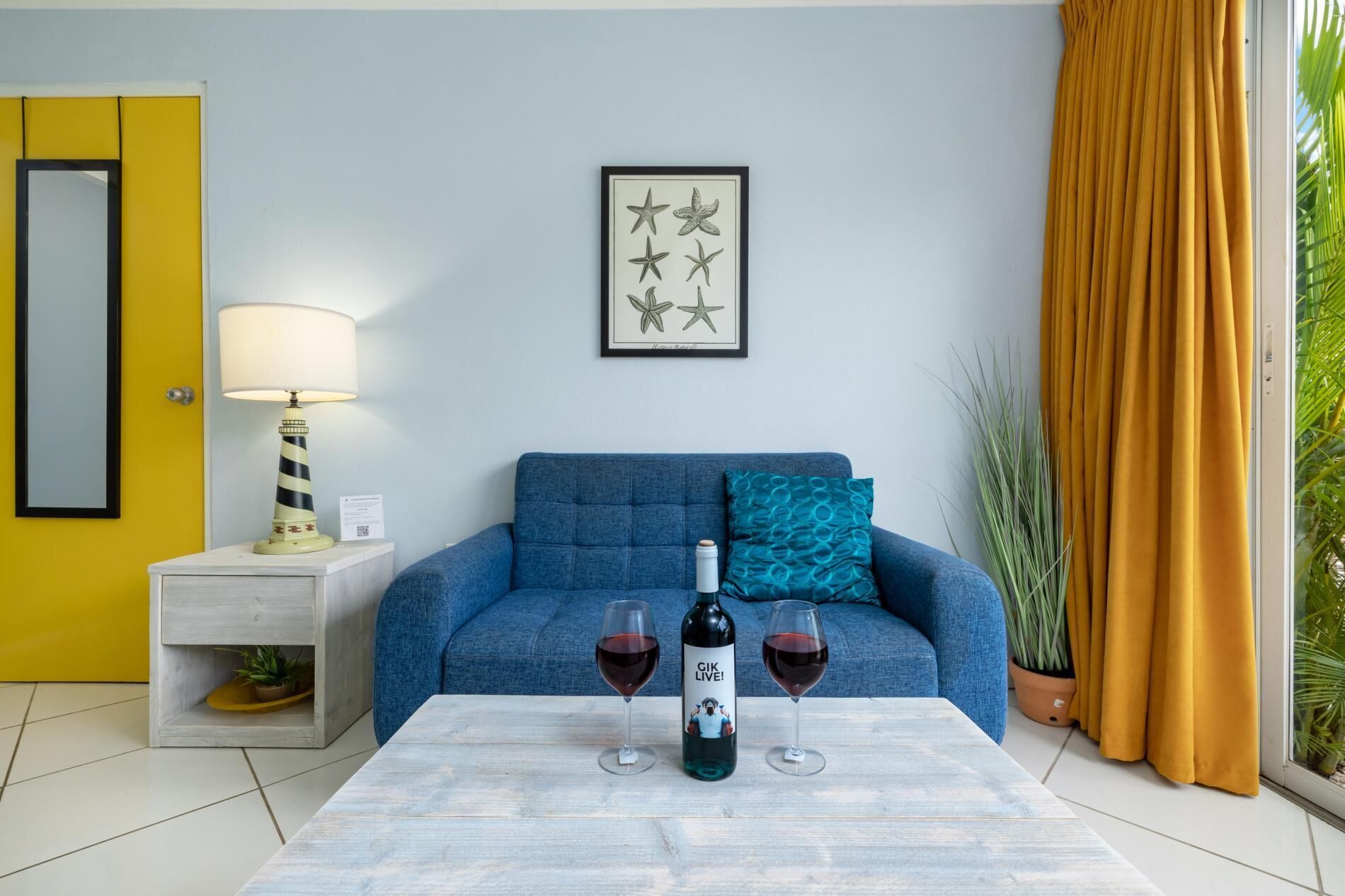 Una stanza con un divano blu, un tavolino con due bicchieri da vino e una bottiglia, una lampada, un quadro incorniciato appeso al muro e una porta gialla.