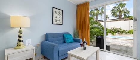A living room with a sofa, a table, a lamp, and a view of a pool and palm trees.