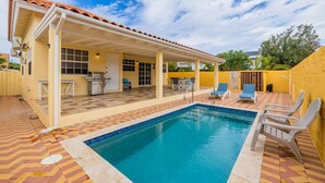 A swimming pool with a tiled deck, a covered patio area with seating, and a yellow exterior wall. - Cute & Cozy 3BR, Great Location (Noord)