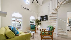 A spacious living area with a green sofa, a wooden chair, and a glass coffee table. A TV is mounted on the wall, and a spiral staircase is visible in the background. - Cute & Cozy 3BR, Great Location (Noord)