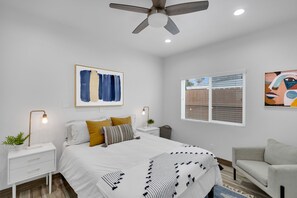 Une chambre à coucher comprenant un lit, des tables de chevet, un ventilateur de plafond, une fenêtre et des œuvres d’art accrochées aux murs.