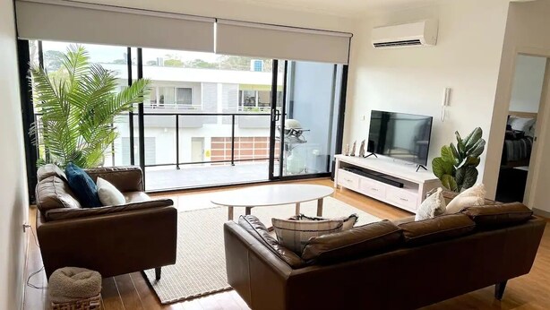 A modern living room with a sofa, a coffee table, and a TV. There is a large window with a view of buildings outside. - Capel Sound Coastal Stays (Capel Sound)