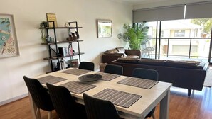 A modern living room with a dining area, featuring a large window, a sofa, a coffee table, and a bookshelf. - Capel Sound Coastal Stays (Capel Sound)