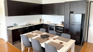 A modern kitchen with dark cabinetry, a stainless steel refrigerator, and a dining table with grey chairs. - Capel Sound Coastal Stays (Capel Sound)