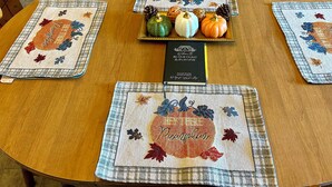 A round wooden table with four placemats featuring autumn-themed embroidery and a small sign.