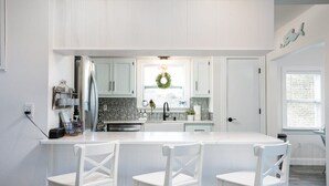 A modern kitchen with white cabinetry, a central island with bar stools, a stainless steel refrigerator, and a window with a view. - Secluded 3min Walk to Beach! Private Pool & Spa (Corolla)