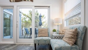 A room with a sliding glass door leading to an outdoor patio, a patterned armchair, a small side table with a lamp, and a view of the outdoors. - Secluded 3min Walk to Beach! Private Pool & Spa (Corolla)