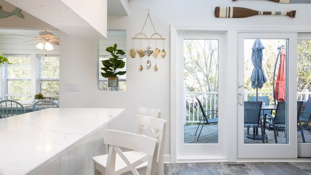 A modern kitchen with a white island and bar stools, a hanging wind chime, and a view of the outdoor deck through glass doors. - Secluded 3min Walk to Beach! Private Pool & Spa (Corolla)