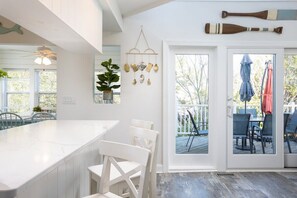 A modern kitchen with a white island and bar stools, a hanging wind chime, and a view of the outdoor deck through glass doors. - Secluded 3min Walk to Beach! Private Pool & Spa (Corolla)