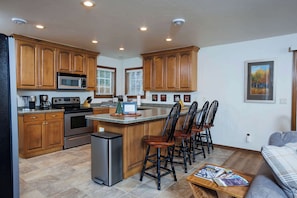 A kitchen with wooden cabinets, a microwave, an oven, and a coffee maker. There are four chairs around a small island, and a sofa in the living area.
