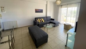 A living room with a sofa, ottoman, glass coffee table, and a dining area with chairs and a table. - Impeccable Apt sleeps 5 in Heraklion (Heraklion)