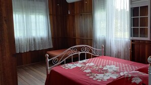 A bedroom with a wooden floor, a bed with a red bedspread, a wooden headboard, and two windows with white curtains. - Poto Mitan (Tonate-Macouria)