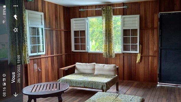 A room with wooden paneling, a bed with a green patterned bedspread, a small table, and two windows with curtains. - Poto Mitan (Tonate-Macouria)