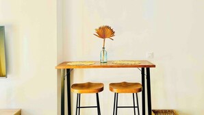 Una pequeña mesa de madera con dos taburetes, un florero con hojas secas y un mantel tejido.