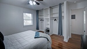 Ein Schlafzimmer mit einem Bett, einem Deckenventilator, einem Fenster mit Jalousien, einem Kleiderschrank mit Regalen und einer Tür.