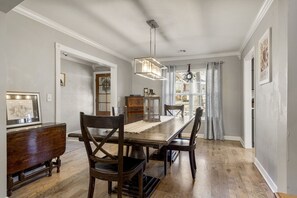 Une salle à manger avec une table et des chaises en bois, un lustre et une vue sur l’extérieur à travers une fenêtre ornée de rideaux.