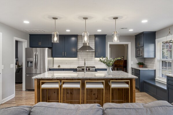 Una cocina moderna con armarios azul oscuro, electrodomésticos de acero inoxidable, una isla central con taburetes de bar, iluminación colgante y vista al área de estar contigua.
